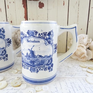 May include: White ceramic beer stein with blue floral and landscape design. The stein features the word "Heineken" and a depiction of a windmill. The handle is a curved shape with blue accents. Buttons and flowers are in the background.