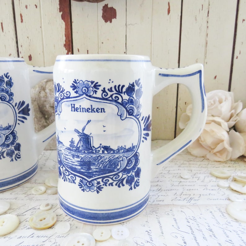 May include: White ceramic beer stein with blue floral and landscape design. The stein features the word "Heineken" and a depiction of a windmill. The handle is a curved shape with blue accents. Buttons and flowers are in the background.