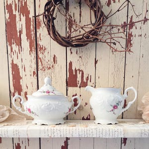 May include: White porcelain sugar bowl with lid and creamer set. Both pieces feature pink rose floral designs and gold trim. The set is displayed on a white shelf with a distressed wood background and a brown twig wreath.
