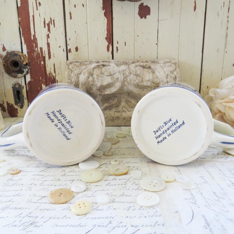 May include: Two white ceramic mugs with blue hand-painted designs. The bottom of each mug features the text "Delfts Blue Handpainted Made in Holland" in blue. The mugs are surrounded by buttons.