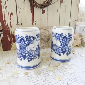 May include: Two white ceramic beer steins with blue floral and windmill designs. Each stein features the word "Heineken" in a decorative banner. The steins have a cylindrical shape with a blue band at the base.