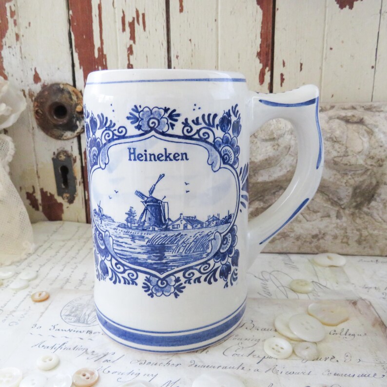 May include: White ceramic beer stein with a blue and white Dutch windmill design. The stein has a handle and the word "Heineken" is displayed above the windmill scene. The design is framed by floral patterns.