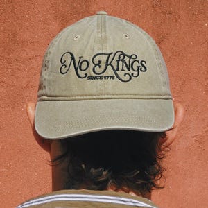 Pode incluir: Bon&eacute; de beisebol verde-oliva com a inscri&ccedil;&atilde;o "No Kings Since 1776" bordada em preto. O bon&eacute; &eacute; usado ao contr&aacute;rio, com as costas do usu&aacute;rio e a camisa listrada vis&iacute;veis em um fundo vermelho-alaranjado.