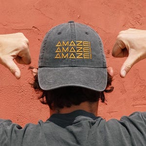 May include: A gray baseball cap with the word "AMAZE" embroidered in yellow three times. The cap is worn backward, with the wearer's back to the camera. Two hands with thumbs down are on either side of the cap.