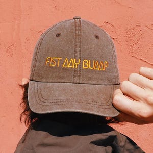 Puede incluir: Gorra de béisbol marrón con el texto "FIST AAY BUUAP" bordado en amarillo. La gorra se lleva hacia atrás, con la visera hacia arriba. El fondo es de color coral sólido.