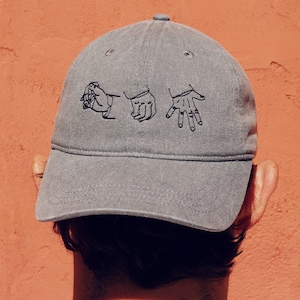 May include: Grey baseball cap with a curved peak. The cap has a black line drawing of three hand gestures. The person wearing the cap is facing away, with a white shirt and a red-orange background.