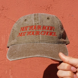 May include: A brown baseball cap with the text "NOT YOUR BODY? NOT YOUR CHOICE." embroidered in red.