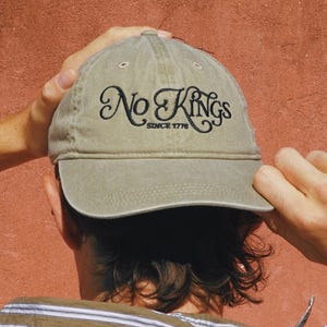 Pode incluir: Bon&eacute; de beisebol c&aacute;qui com o texto bordado "No Kings Since 1776" em escrita preta. O bon&eacute; est&aacute; sendo segurado por uma pessoa vestindo uma camisa listrada contra um fundo vermelho-alaranjado.