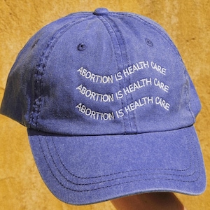 May include: A blue denim baseball cap with the phrase "Abortion is Health Care" embroidered in white on the front.