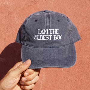 May include: A charcoal grey baseball cap with a washed-out finish featuring the white embroidered text I AM THE ELDEST BOY centered on the front panel. The cap has a curved brim, a button on the top, and stitched eyelets for ventilation.
