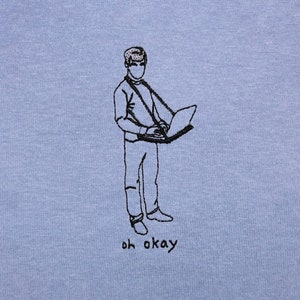 Pode incluir: Camiseta azul clara com um desenho a traço preto de uma pessoa segurando um laptop. O texto "oh okay" está impresso em preto abaixo. A camiseta tem gola redonda e mangas curtas.