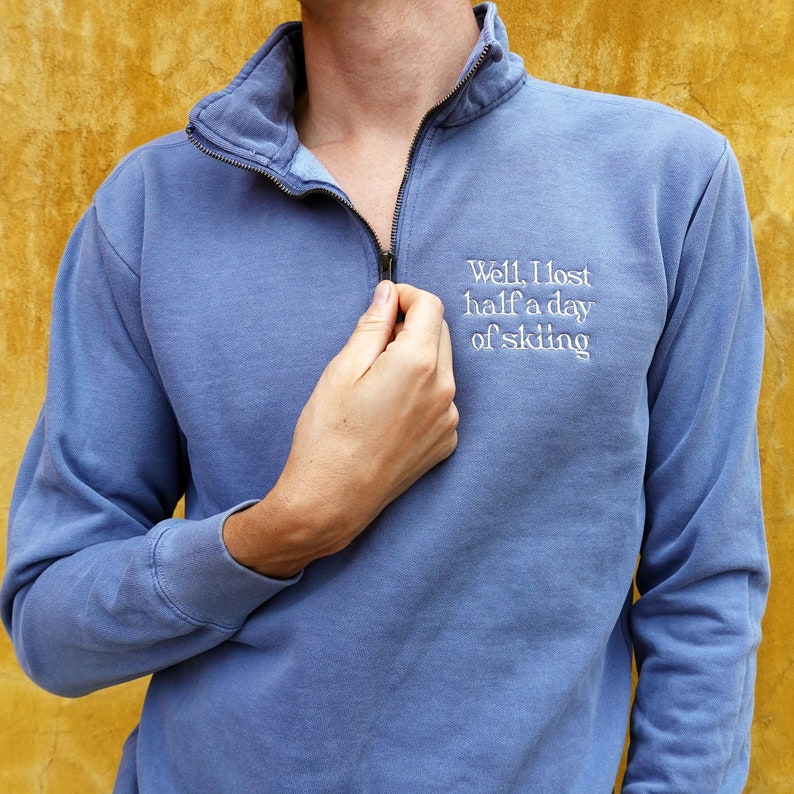 K&ouml;nnte beinhalten: Blaues Sweatshirt mit Rei&szlig;verschluss am Kragen. Auf dem Sweatshirt steht in Wei&szlig; gestickt: "Well, I lost half a day of skiing". Das Model tr&auml;gt das Sweatshirt.