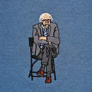 Könnte beinhalten: Gestickte Abbildung einer Person, die auf einem Stuhl sitzt. Die Person trägt einen grauen Anzug und eine blaue Krawatte. Der Stuhl ist schwarz und hat ein schlichtes Design.