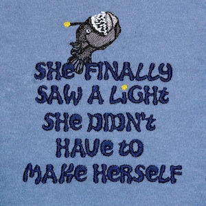 May include: Blue t-shirt with an embroidered anglerfish and the text "She finally saw a light she didn't have to make herself."