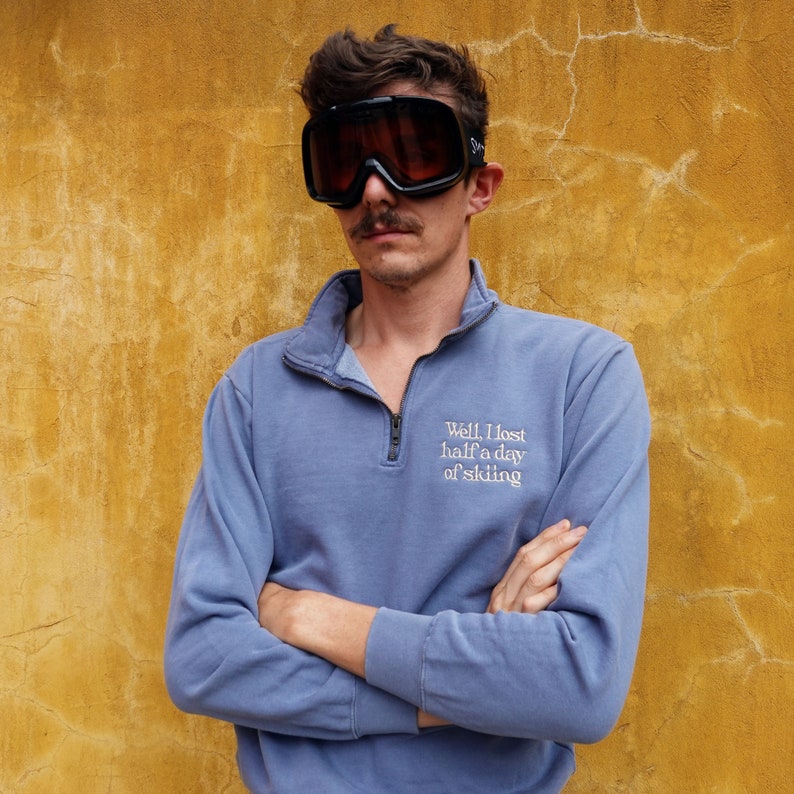 K&ouml;nnte beinhalten: Eine Person tr&auml;gt eine schwarze Skibrille und einen blauen Pullover mit einem Kragen mit Viertelrei&szlig;verschluss. Der Pullover hat den gestickten Text "Well, I lost half a day of skiing". Der Hintergrund ist eine strukturierte gelbe Wand.