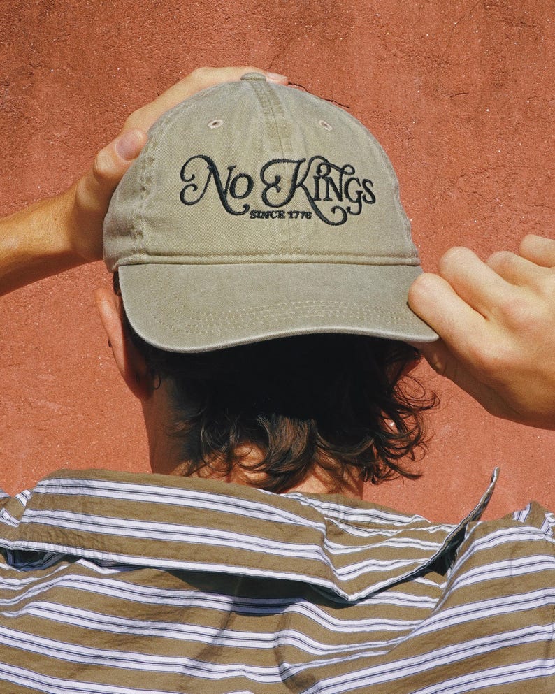 Pode incluir: Bon&eacute; de beisebol c&aacute;qui com o texto bordado "No Kings Since 1776" em escrita preta. O bon&eacute; est&aacute; sendo segurado por uma pessoa vestindo uma camisa listrada contra um fundo vermelho-alaranjado.