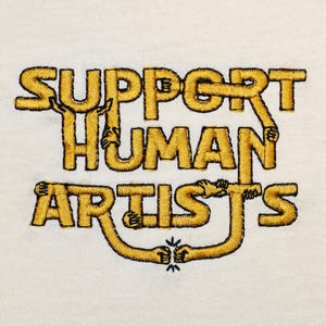 Może przedstawiać: Biały t-shirt z haftem w kolorze złotym, na którym widnieje napis "Support Human Artists". Tekst tworzą stylizowane dłonie trzymające się nawzajem.
