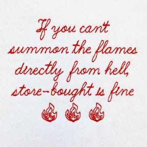 Pode incluir: Texto bordado em vermelho sobre um fundo branco que diz "Se você não pode invocar as chamas diretamente do inferno, as da loja estão bem." Três ilustrações de chamas vermelhas estão abaixo do texto.