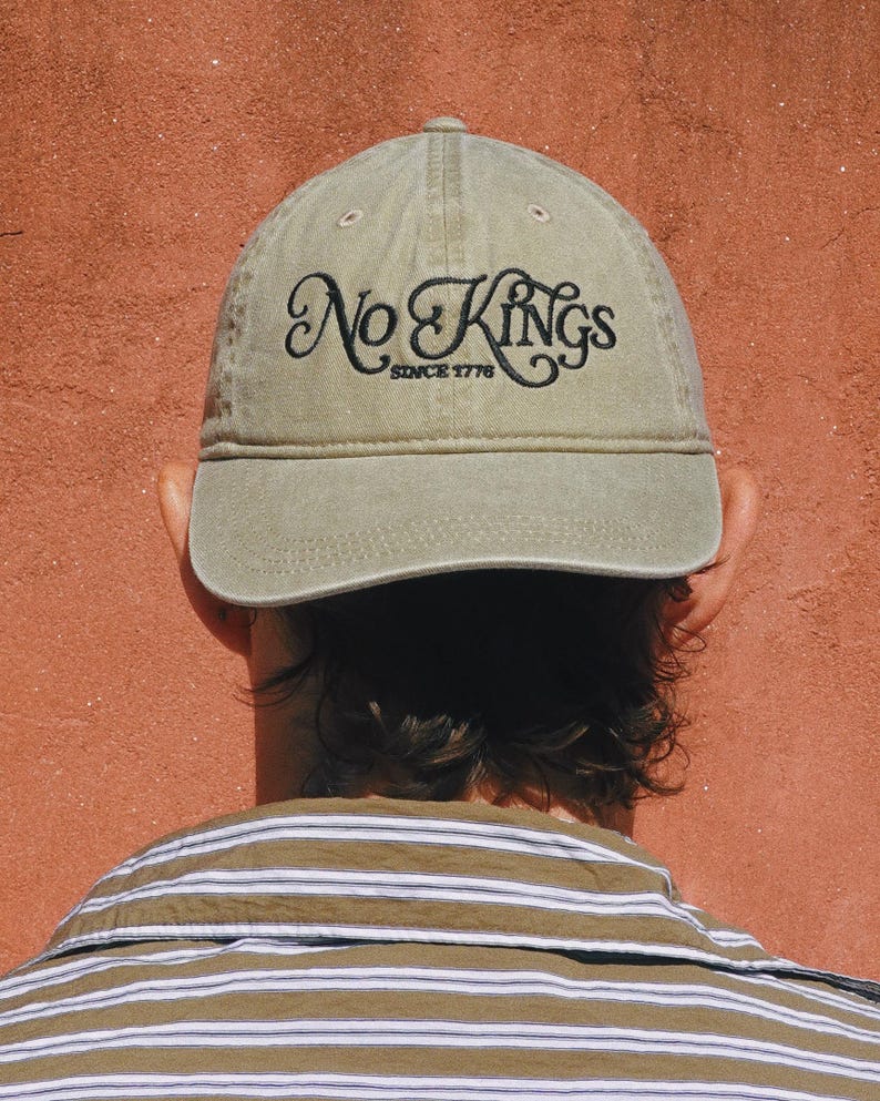 Pode incluir: Bon&eacute; de beisebol verde-oliva com a inscri&ccedil;&atilde;o "No Kings Since 1776" bordada em preto. O bon&eacute; &eacute; usado ao contr&aacute;rio, com as costas do usu&aacute;rio e a camisa listrada vis&iacute;veis em um fundo vermelho-alaranjado.
