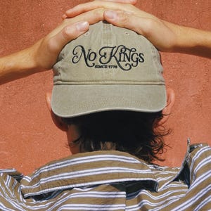 Pode incluir: Bon&eacute; de beisebol c&aacute;qui com o texto "No Kings Since 1776" bordado em escrita preta. O bon&eacute; &eacute; usado por uma pessoa que usa uma camisa listrada marrom e branca. O fundo &eacute; de cor vermelho-alaranjado s&oacute;lido.
