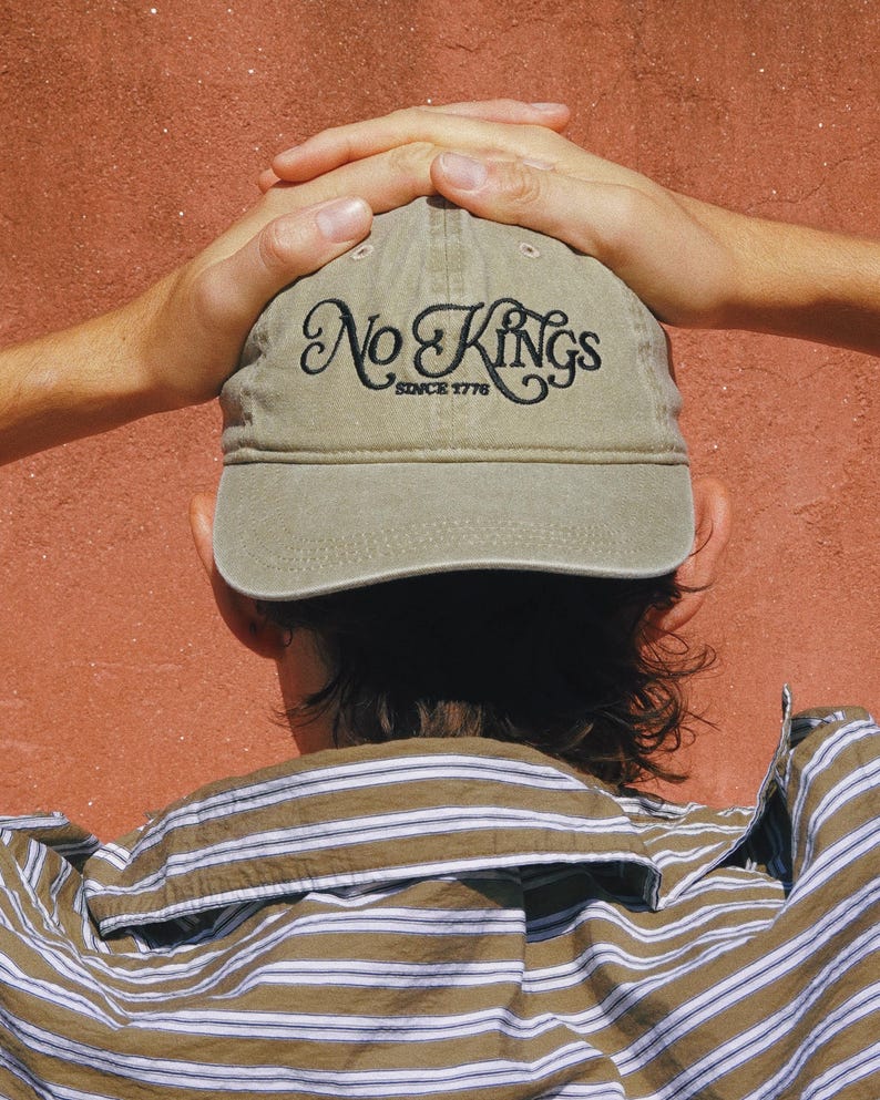 Pode incluir: Bon&eacute; de beisebol c&aacute;qui com o texto "No Kings Since 1776" bordado em escrita preta. O bon&eacute; &eacute; usado por uma pessoa que usa uma camisa listrada marrom e branca. O fundo &eacute; de cor vermelho-alaranjado s&oacute;lido.