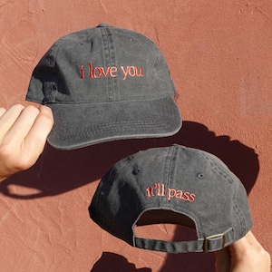 May include: A charcoal grey baseball cap featuring the embroidered red text i love you on the front panel and the embroidered red text it'll pass on the back panel. The cap includes a curved brim, a six-panel construction, and an adjustable strap.