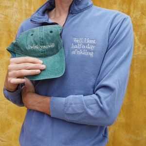 K&ouml;nnte beinhalten: Ein blaues Langarm-Pullover mit einem Viertel-Rei&szlig;verschlusskragen und dem gestickten Text "Well, I lost half a day of skiing." Eine gr&uuml;ne Baseballkappe mit dem Text "I wish you well" wird vor dem Pullover gehalten.