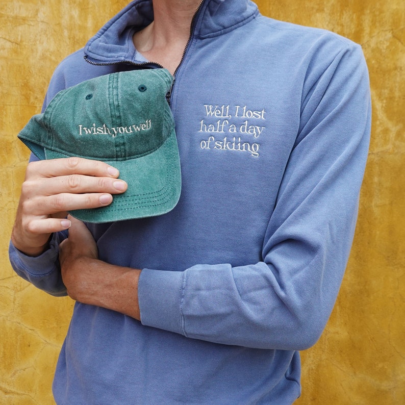 K&ouml;nnte beinhalten: Ein blaues Langarm-Pullover mit einem Viertel-Rei&szlig;verschlusskragen und dem gestickten Text "Well, I lost half a day of skiing." Eine gr&uuml;ne Baseballkappe mit dem Text "I wish you well" wird vor dem Pullover gehalten.