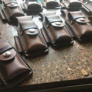 May include: Several handcrafted brown leather pouches are displayed. Each pouch features a snap closure, a small side pocket, and is stitched with white thread. The dark brown leather strap and the leather's textured appearance are visible.