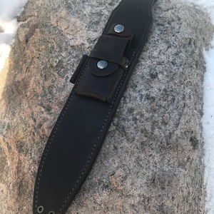May include: A black leather knife sheath with a belt loop and snap closures. The sheath has a stitched border and a small pouch. The background is a gray rock with patches of snow.