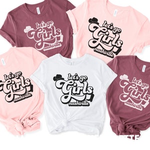 May include: Five t-shirts with the text "Let's go Girls Nashville" in a retro font. The shirts are in pink, white, and maroon.