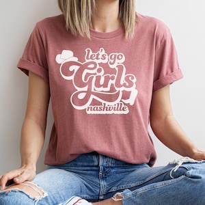 May include: A mauve t-shirt with a white graphic that says "Let's go girls Nashville" in a retro font.