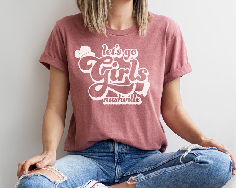 May include: A mauve t-shirt with a white graphic that says "Let's go girls Nashville" in a retro font.