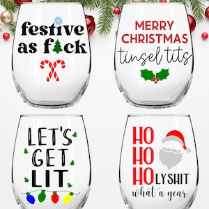 May include: Four stemless wine glasses with humorous Christmas-themed text printed on them. The glasses feature the phrases "festive as f*ck", "Merry Christmas tinsel tits", "Let's get lit", and "Ho Ho Ho LYSHIT what a year".