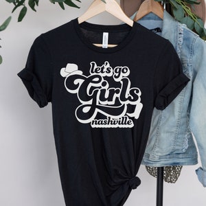 May include: Black t-shirt with white text that says "Let's go girls Nashville" with a small cowboy hat graphic above the text.