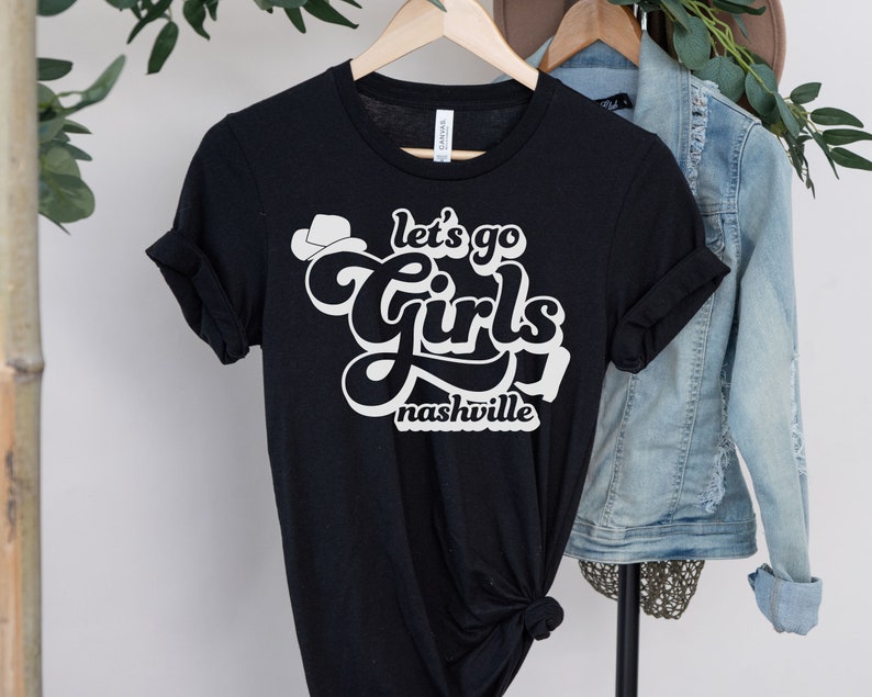 May include: Black t-shirt with white text that says "Let's go girls Nashville" with a small cowboy hat graphic above the text.