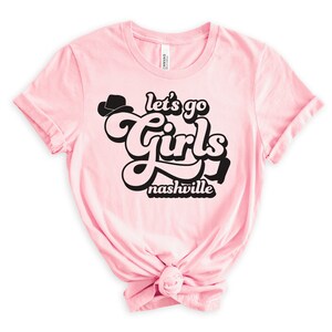 May include: Pink t-shirt with a black graphic that says "Let's go girls Nashville" in a retro font with a cowboy hat graphic.