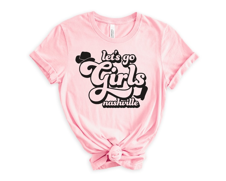 May include: Pink t-shirt with a black graphic that says "Let's go girls Nashville" in a retro font with a cowboy hat graphic.