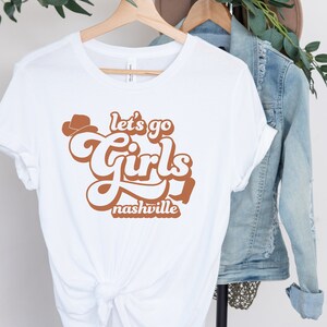 May include: White t-shirt with a brown graphic that says "Let's go girls Nashville" in a retro font.