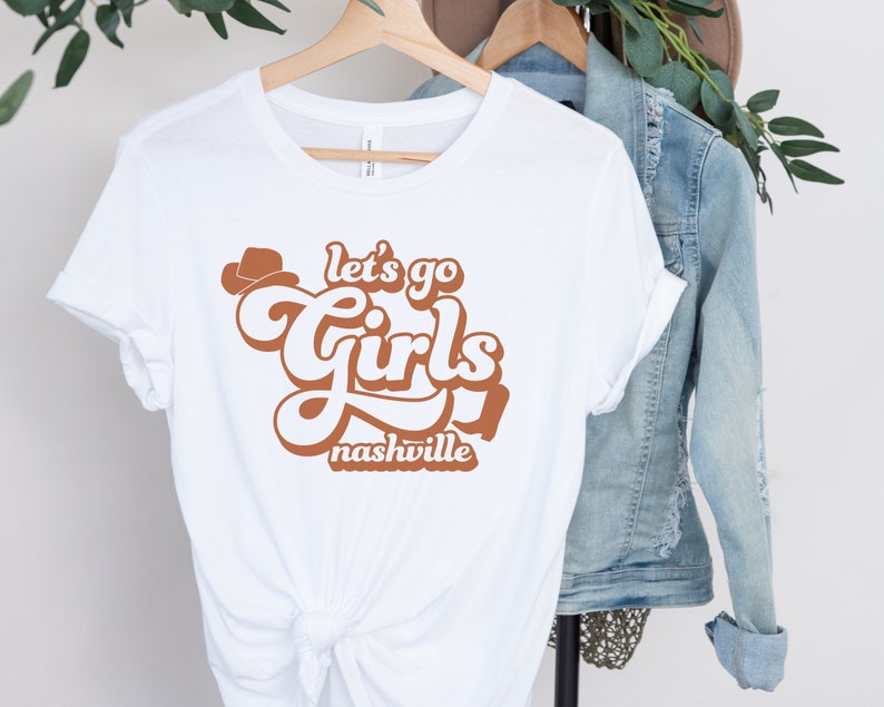 May include: White t-shirt with a brown graphic that says "Let's go girls Nashville" in a retro font.
