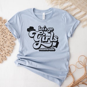 May include: Light blue t-shirt with black text that reads "Let's go girls Nashville" in a retro font. A black cowboy hat is above the word "girls".