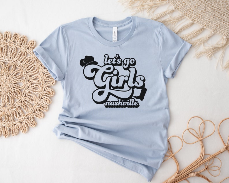 May include: Light blue t-shirt with black text that reads "Let's go girls Nashville" in a retro font. A black cowboy hat is above the word "girls".