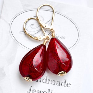 Peut inclure: Une paire de boucles d'oreilles à fermoir levier doré avec des perles en verre rouge profond en forme de goutte et une texture marbrée. Les boucles d'oreilles sont présentées sur une carte blanche portant l'inscription "Handmade Jewellery".