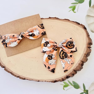 May include: Two hair bows made of light orange fabric featuring a repeating pattern of Winnie the Pooh characters and black jack-o'-lanterns. One bow is attached to a rectangular brown card, while the other is a larger, multi-layered bow.