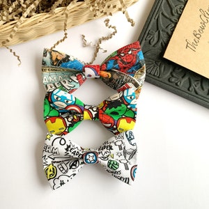 May include: Three bow ties featuring different superhero comic book designs. The top bow tie has a red, blue and yellow pattern with Spiderman, Captain America and Iron Man. The middle bow tie has a green, red and yellow pattern with Captain America, Iron Man and Hulk. The bottom bow tie has a white background with black, red, blue and green comic book characters and text.