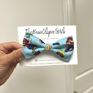 Puede incluir: Una pajarita azul claro con un patrón repetido de personajes de Mario del videojuego Super Mario Bros. La pajarita tiene un nudo central con un diseño de bloque amarillo. El logotipo de The Bow Clique Girls es visible en la tarjeta adjunta.