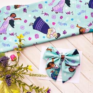 May include: A turquoise blue fabric bow with a print of a cartoon character from the movie Encanto. The bow is on a white card with the text "The Bowtique" printed on it.