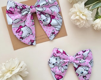 May include: Two fabric hair bows featuring a repeating pattern of white cartoon kittens with pink bows on a light pink background with darker pink polka dots. The bows are constructed with a central knot and have pointed ends on each side.