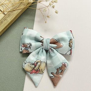 May include: A light blue plaid fabric bow featuring illustrations of Winnie the Pooh characters. The bow is tied in the center and has pointed ends. The background is white and light green.