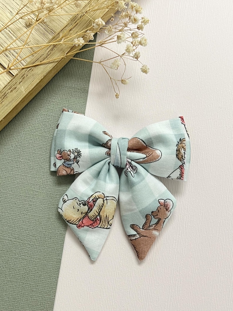 May include: A light blue plaid fabric bow featuring illustrations of Winnie the Pooh characters. The bow is tied in the center and has pointed ends. The background is white and light green.